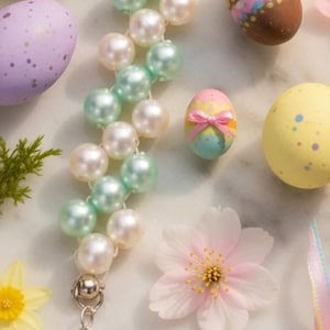 May include: An Easter-themed image featuring a pearl bracelet with pastel green and white beads, surrounded by decorated Easter eggs in various colors, a small chick, and flowers. The bracelet has a heart-shaped charm.