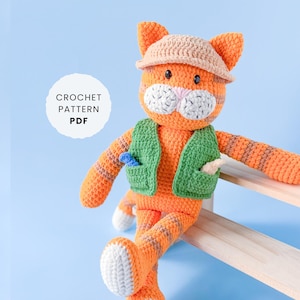 May include: A crocheted orange cat doll with brown stripes, wearing a green waistcoat and a beige hat. The cat has white accents on its face and feet. The image includes the text "CROCHET PATTERN PDF" in a white circle.