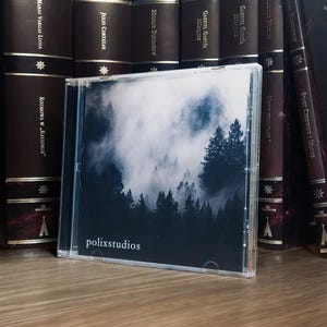 May include: A CD case displaying a forest scene shrouded in mist, with the text "polixstudios". The CD is positioned in front of a row of hardback books with gold lettering and detailing.