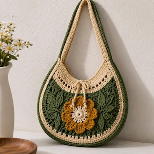 Könnte beinhalten: Eine halbmondförmige Häkeltasche mit dunkelgrünem Körper, cremefarbenem Rand und einem einzelnen Schulterriemen. Die Vorderseite ist mit einer großen, strukturierten senfgelben Blumenapplikation, zwei grünen Blattmotiven und einem Kordelzug verziert.