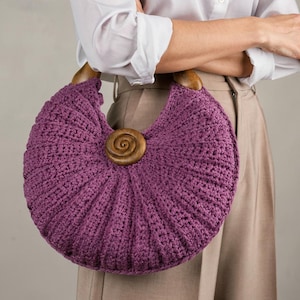 Puede incluir: Un bolso de ganchillo morado en forma de media luna con un patr&oacute;n acanalado texturizado, dos asas semicirculares de madera marr&oacute;n oscuro y una pieza central de madera con forma de espiral fijada en la parte frontal.