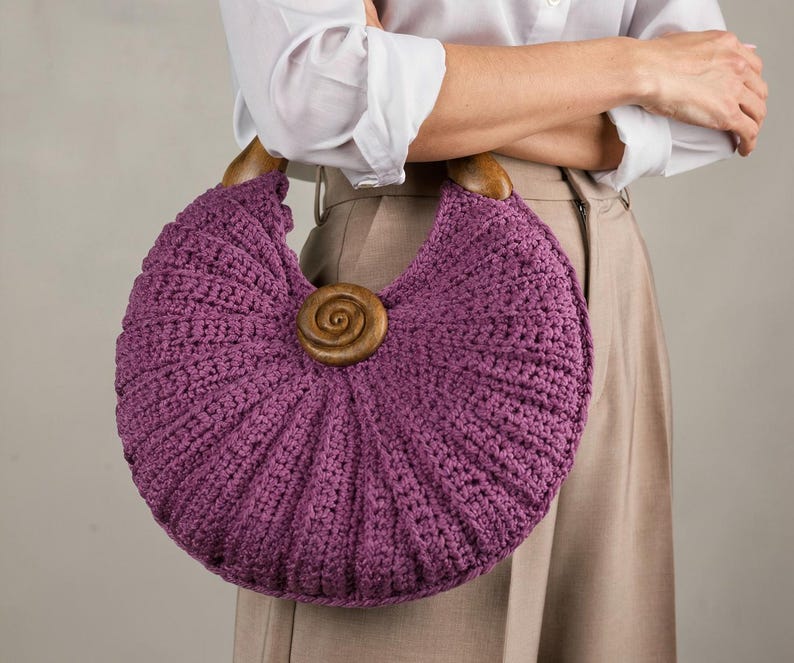 Puede incluir: Un bolso de ganchillo morado en forma de media luna con un patr&oacute;n acanalado texturizado, dos asas semicirculares de madera marr&oacute;n oscuro y una pieza central de madera con forma de espiral fijada en la parte frontal.
