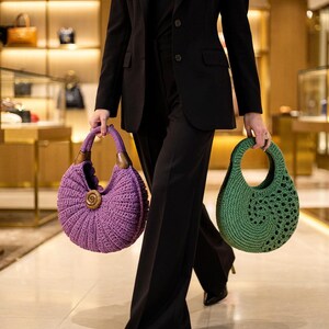 Puede incluir: Un bolso de ganchillo morado con asa de madera y detalle en espiral junto a un bolso de ganchillo verde con asa circular recortada. Ambos bolsos tienen una forma redondeada de media luna y son sostenidos por una persona con traje negro.