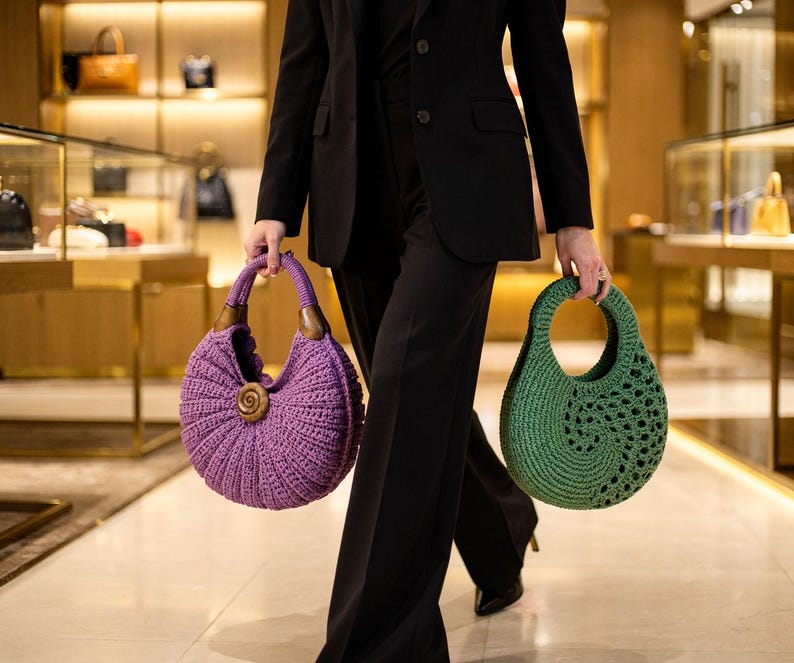 Puede incluir: Un bolso de ganchillo morado con asa de madera y detalle en espiral junto a un bolso de ganchillo verde con asa circular recortada. Ambos bolsos tienen una forma redondeada de media luna y son sostenidos por una persona con traje negro.