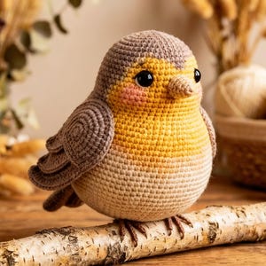 Könnte beinhalten: Ein gehäkeltes Vogelspielzeug mit gelber Brust, beigem Körper und braunen Flügeln und Kopf. Der Vogel hat schwarze Augen und sitzt auf einem Birkenast. Der Hintergrund zeigt getrocknete Blumen und Wollknäuel.