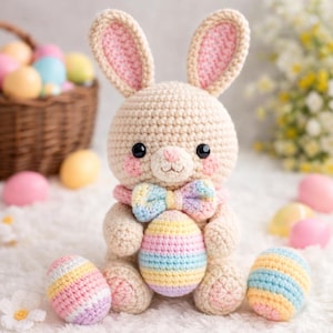 Könnte beinhalten: Ein gehäkelter Hase mit pastellfarbener Fliege und rosigen Wangen sitzt neben drei gehäkelten Ostereiern. Der Hase hält ein gestreiftes Ei. Ein Korb mit bunten Eiern befindet sich im Hintergrund. Die Ohren des Hasen sind rosa gefüttert.