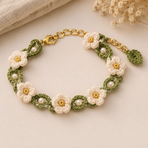 Könnte beinhalten: Ein Armband mit gehäkelten cremefarbenen Blumen mit gelben Zentren, durchsetzt mit kleinen weißen Perlen und grünen gehäkelten blattförmigen Gliedern. Das Schmuckstück enthält eine goldfarbene Metallkette, einen Karabinerverschluss und einen kleinen grünen Blattanhänger.