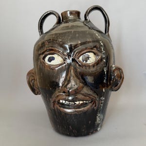 May include: A dark brown glazed ceramic face jug featuring two arched handles at the top, a sculpted human face with wide eyes, a prominent nose, and a mouth showing white teeth. The glossy finish has subtle variations in tone and texture.