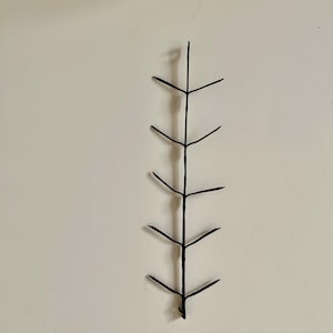 May include: A vertical black metal wall sculpture shaped like a slender branch or stem. The object features a central spine with five pairs of thin, symmetrical, outward-extending twigs or leaves arranged in an alternating pattern along the length.