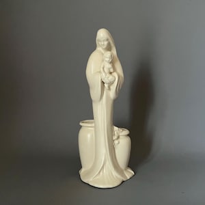 May include: A cream-colored ceramic statue of a woman holding a child. The woman is draped in flowing robes, and the statue has a vase-like base. The sculpture is a religious figure, likely a depiction of the Virgin Mary and Jesus.