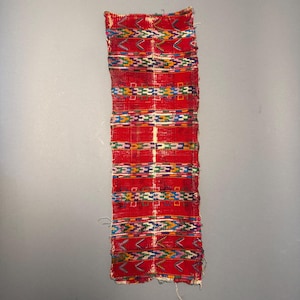 May include: A rectangular textile strip featuring a vibrant red base color with horizontal bands of intricate geometric patterns in blue, yellow, green, and white. The fabric displays a woven texture with frayed edges at the top and bottom ends.