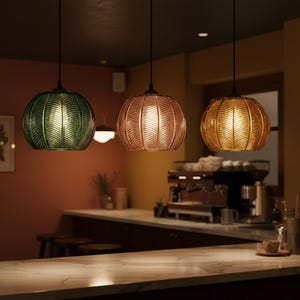 May include: Three pendant lights with textured, spherical glass shades in green, copper, and gold. Each light hangs from a black cord, illuminating a bar area with a marble countertop and a coffee machine.
