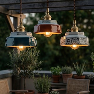 May include: Three pendant lights with ceramic shades in teal, rust, and white, each with a brass fixture and exposed bulb. The lights hang from brown cords against a blurred outdoor background, suggesting a patio or deck setting. The lights are a decorative lighting fixture.