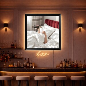 May include: A black framed wall art with a backlit border. The art shows a person pouring champagne into a glass while in bed. The word "bar" is written in neon lights below the art.