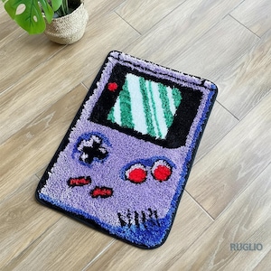 May include: A purple and black rug shaped like a retro handheld video game console. The rug features a screen with green and white stripes, red buttons, and a black directional pad. The rug is placed on a wooden floor.