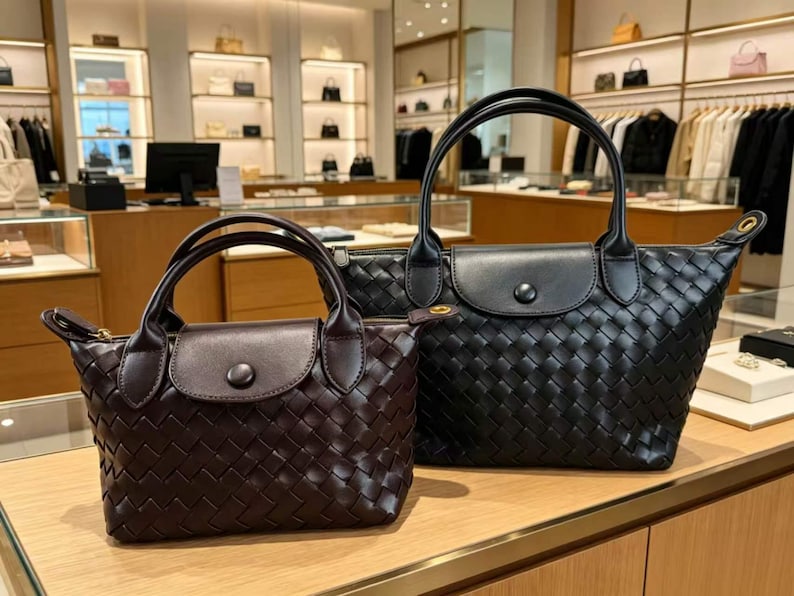 May include: Two woven handbags with top handles and a front flap closure featuring a single button. The smaller bag is dark brown, while the larger bag is black. Both bags have a distinct woven texture across the body and smooth leather accents.