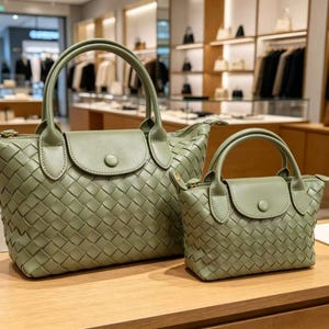 May include: Two sage green handbags featuring a woven exterior pattern, dual top handles, and a front flap closure with a single button. The bags are structured with a smooth finish on the flap and handles, showcasing a consistent design in two sizes.