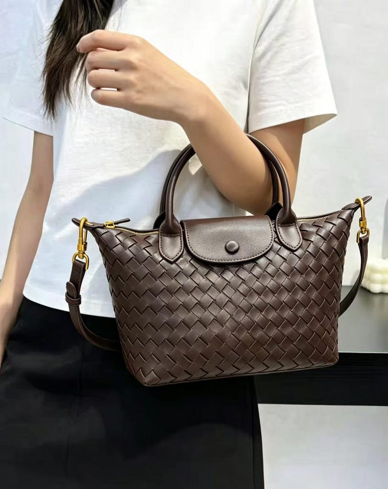 May include: A dark brown handbag featuring a woven exterior pattern, two top handles, and a central flap with a snap button closure. The bag includes a detachable shoulder strap with gold-toned metal hardware and a zippered top opening.