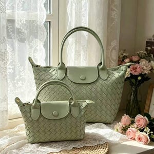 May include: Two sage green handbags featuring a woven texture, dual top handles, and a front flap closure with a single button snap. The larger bag is positioned behind the smaller bag, both displaying a consistent light green color and pattern.
