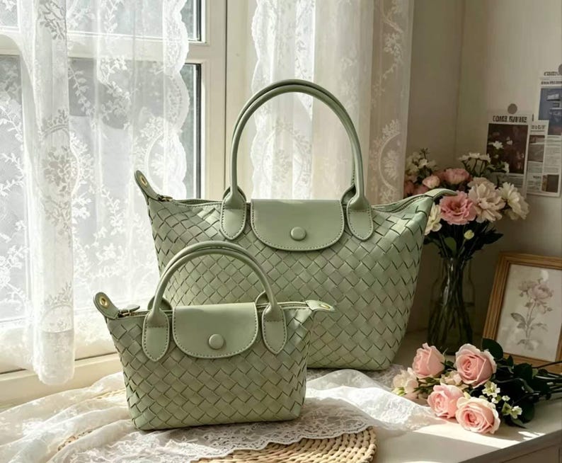 May include: Two sage green handbags featuring a woven texture, dual top handles, and a front flap closure with a single button snap. The larger bag is positioned behind the smaller bag, both displaying a consistent light green color and pattern.
