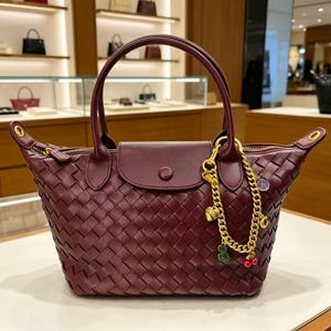 May include: A deep burgundy handbag featuring a woven leather exterior, two top handles, and a central flap with a snap closure. A gold-toned chain charm with small colorful fruit and heart-shaped pendants is attached to one of the handle bases.