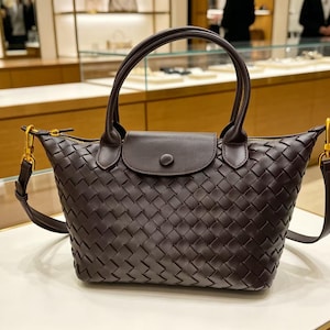 May include: A dark brown handbag featuring an intricate woven leather pattern across the body. The bag includes two rounded top handles, a central flap with a single snap button closure, and gold-toned metal hardware attachments for a shoulder strap.