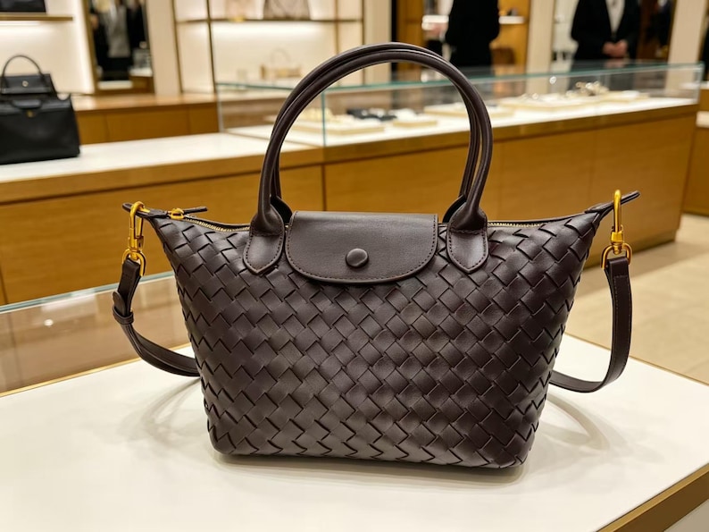 May include: A dark brown handbag featuring an intricate woven leather pattern across the body. The bag includes two rounded top handles, a central flap with a single snap button closure, and gold-toned metal hardware attachments for a shoulder strap.