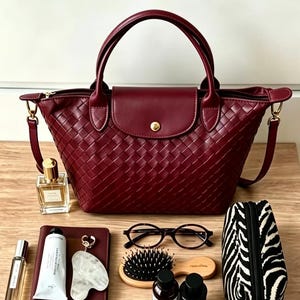 May include: A deep burgundy woven handbag with two top handles, a central flap featuring a gold-tone snap closure, and side attachments for a shoulder strap. The bag is surrounded by a perfume bottle, a notebook, a tube, a stone tool, glasses, a hairbrush, two small bottles, and a zebra-print pouch.