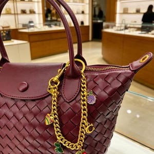 May include: A deep burgundy woven leather handbag featuring a gold-tone chain bag charm. The charm includes a striped heart, a purple flower, a gold letter C, a green dinosaur, and red cherries. The bag has a top handle and a snap-button flap.
