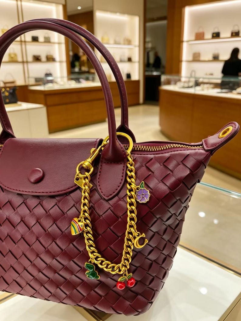 May include: A deep burgundy woven leather handbag featuring a gold-tone chain bag charm. The charm includes a striped heart, a purple flower, a gold letter C, a green dinosaur, and red cherries. The bag has a top handle and a snap-button flap.