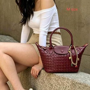 May include: A burgundy handbag with a woven design, featuring a top handle and a flap closure. A gold chain with charms hangs from the handle. The bag is sitting on a stone surface. The text "M size" is visible.