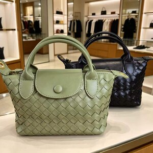 May include: A sage green woven handbag with a top handle, a front flap featuring a single button closure, and gold-tone hardware accents. Positioned behind it is a matching black woven handbag with a similar top handle and front flap design.