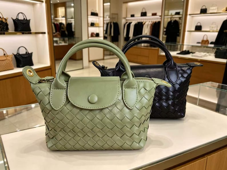May include: A sage green woven handbag with a top handle, a front flap featuring a single button closure, and gold-tone hardware accents. Positioned behind it is a matching black woven handbag with a similar top handle and front flap design.
