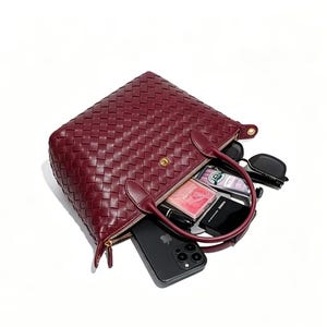 May include: A deep burgundy woven leather handbag with two top handles and a gold-tone snap closure. The bag is open, revealing contents including a black smartphone, a small black compact, a pink rectangular case, a tube, and black sunglasses.