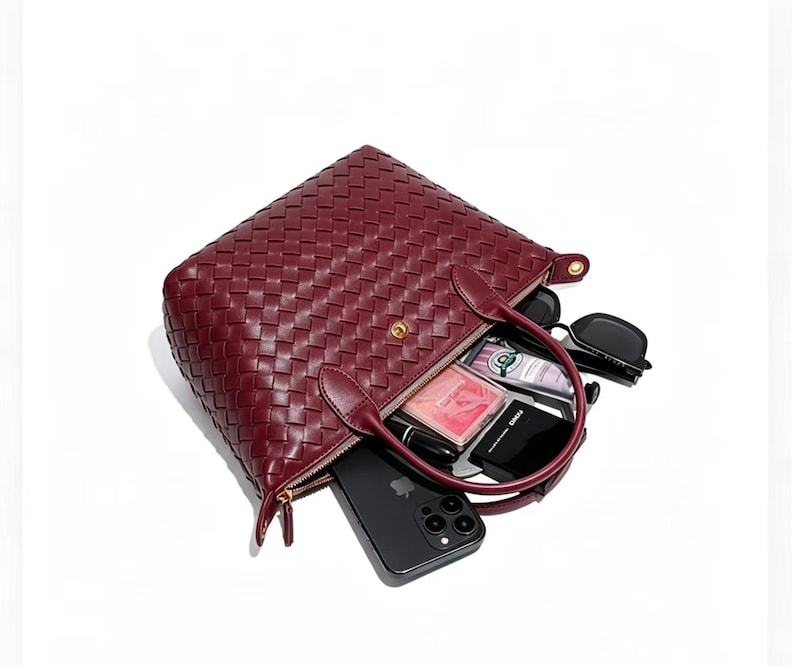 May include: A deep burgundy woven leather handbag with two top handles and a gold-tone snap closure. The bag is open, revealing contents including a black smartphone, a small black compact, a pink rectangular case, a tube, and black sunglasses.