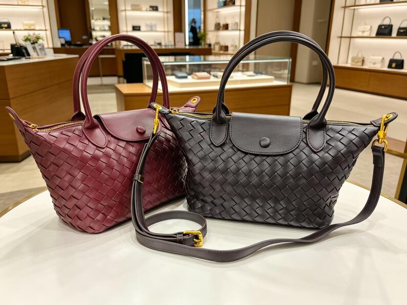 May include: Two woven handbags, one in deep burgundy and one in black, featuring a top handle, a fold-over flap with a snap button closure, a gold-tone zipper, and a detachable, adjustable shoulder strap with gold-tone hardware and a buckle.