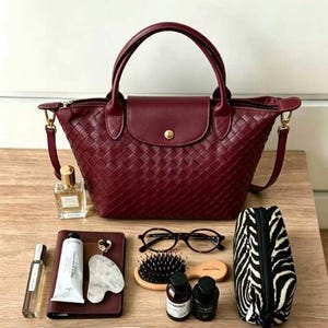 May include: A deep burgundy woven handbag with a flap closure and gold-tone hardware. Surrounding items include a perfume bottle, a notebook, a tube of cream, a keychain, eyeglasses, a hairbrush, two small bottles, and a black and white zebra print pouch.