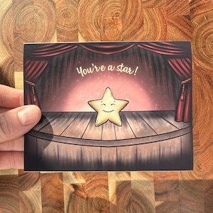 May include: A greeting card featuring a cartoon star on a stage with red curtains. The card has the text "You're a star!" written in a script font. The star has a smiling face.