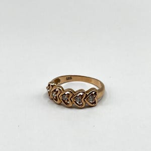 Könnte beinhalten: Ein goldener Ring mit fünf herzförmigen Fassungen, die jeweils einen kleinen, klaren Edelstein halten. Der Ring ist vor einem schlichten weißen Hintergrund platziert, der sein zartes Design und das Funkeln der Steine hervorhebt.