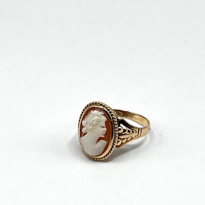 Könnte beinhalten: Ein goldener Ring mit einem ovalen Cameo, der das Profil einer Frau zeigt. Der Cameo ist weiß auf einem rotbraunen Hintergrund, eingefasst in Gold mit einem seilartigen Rand. Der Ring hat ein dekoratives Band.