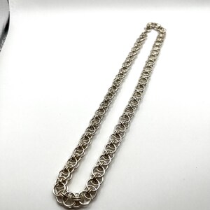 May include: A silver-colored chain necklace with a series of interlocking circular links. The necklace is displayed against a white background, highlighting its metallic sheen and intricate design. The chain is a classic jewelry piece.