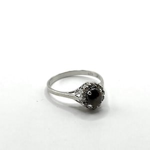 Könnte beinhalten: Ein silberner Ring mit einem dunklen Edelstein, umgeben von kleineren Steinen. Der Ring hat ein zartes Band und eine Halo-Fassung, die ein klassisches und elegantes Design schafft. Der Ring ist vor einem weißen Hintergrund platziert.