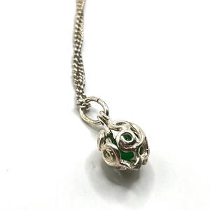 Può includere: Una collana d'argento con un ciondolo dal design traforato. Il ciondolo racchiude una piccola gemma verde e rotonda. La catena è in un delicato stile intrecciato e la chiusura è visibile in alto. La collana è presentata su sfondo bianco.