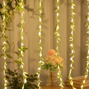 Op de afbeelding: Een reeks verticaal hangende lichtsnoeren met kleine groene kunstbladeren afgewisseld met warmwitte LED-lampjes. De snoeren zijn parallel gerangschikt, wat een gordijneffect creëert met een warme, gloeiende verlichting over het geheel.