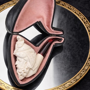 Puede incluir: Una pipa de tabaco tallada de color marfil con un busto detallado de un hombre con casco, exhibida en un estuche de cuero negro con interior rosa. La pipa tiene una boquilla de color rojo oscuro. El estuche está sobre un espejo con borde dorado.