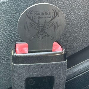 Könnte beinhalten: Ein Metalleinsatz für den Sicherheitsgurt mit dem Text "Natürlich Grünewald" und einem Hirschkopf-Design. Der Einsatz wird in eine schwarze Sicherheitsgurtschnalle eingesetzt. Die Schnalle hat rote Entriegelungsknöpfe.