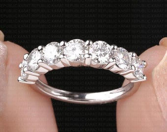 May include: A silver ring featuring a row of seven round, clear gemstones. The ring is held between two fingers against a black background with repeating text.