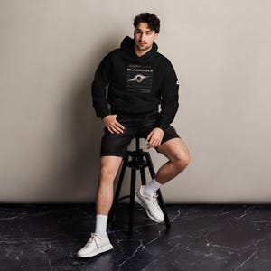 Puede incluir: Una sudadera negra con capucha que presenta un estampado gráfico de un agujero negro en el pecho. El diseño incluye el texto "BLACKHOLE" en letras mayúsculas blancas, rodeado de elementos de texto más pequeños y una ilustración central.