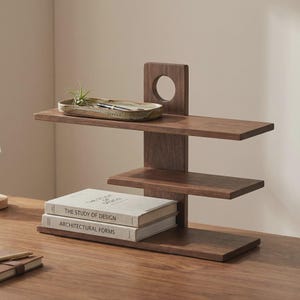 May include: A dark brown wooden desk organizer with three shelves and a vertical support with a circular cutout. Two books with the titles "THE STUDY OF DESIGN" and "ARCHITECTURAL FORMS" are on the bottom shelf.