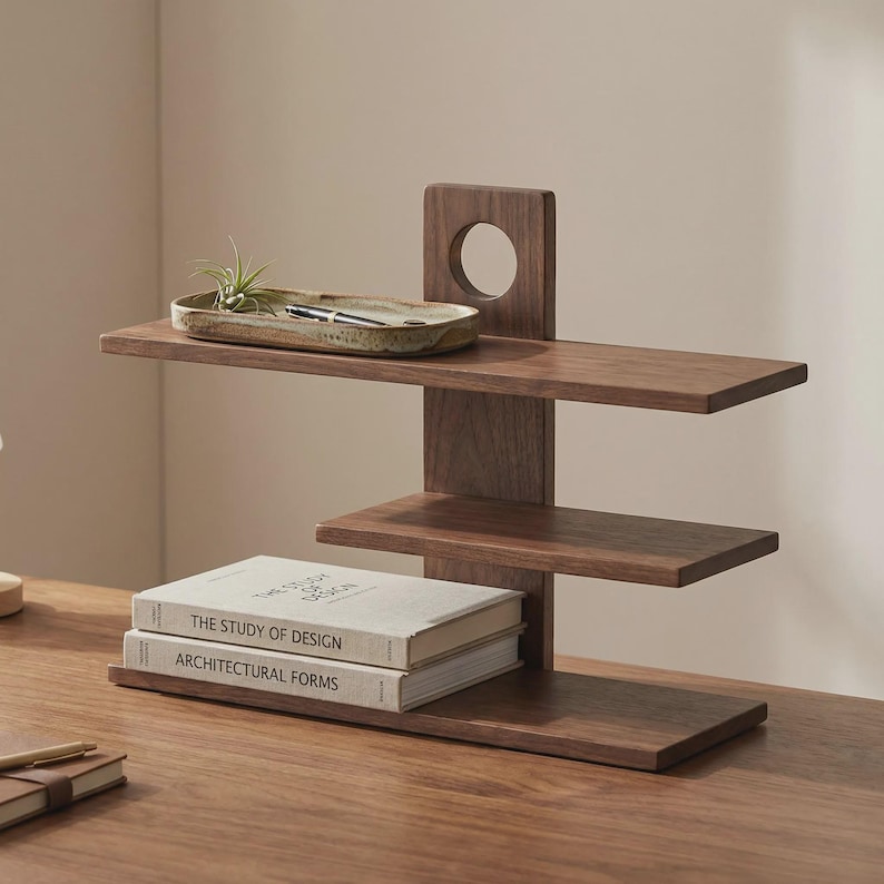 May include: A dark brown wooden desk organizer with three shelves and a vertical support with a circular cutout. Two books with the titles "THE STUDY OF DESIGN" and "ARCHITECTURAL FORMS" are on the bottom shelf.